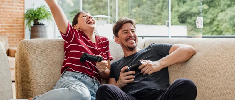 A man and woman are sitting on a light-colored sofa in a sunlit living room with large glass windows, laughing and holding video game controllers, with the woman raising her arm in celebration.